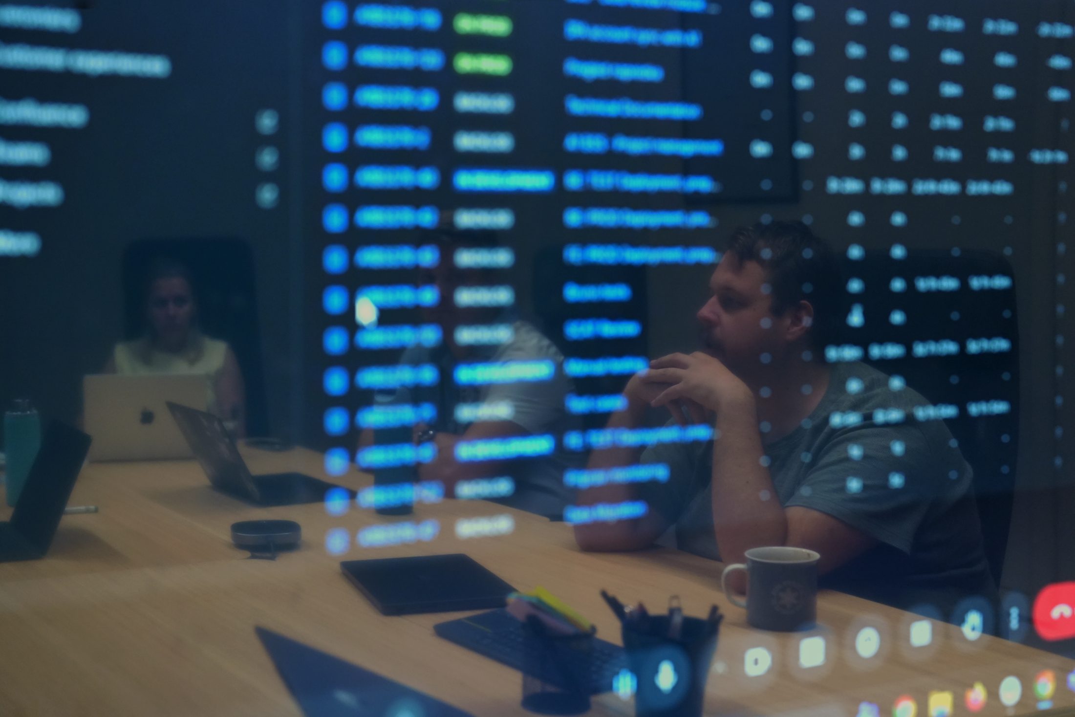 U-centrix and A1 team members working together around a meeting-room table with laptops, with a large screen reflected in the glass